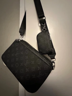 Louis Vuitton Väska  - Snygg svart axelväska från Louis Vuitton med klassiskt monogrammönster i grått. Väskan har bred justerbar axelrem och en liten avtagbar ficka framtill. Tillverkad i tåligt lädermaterial och har silverfärgade detaljer. Perfekt för dig som vill ha en trendig och praktisk väska.