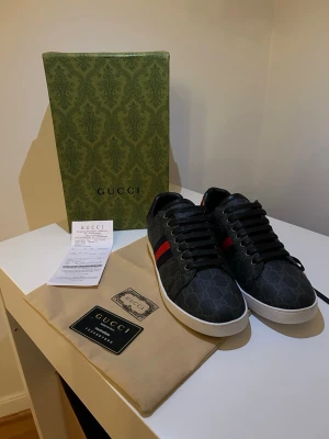 Gucci GG Supreme sneakers svart/röd - Snygga Gucci sneakers i svart GG Supreme canvas med klassiska röda och blå ränder på sidan. Skorna har vita sulor, svarta skosnören och röd detalj på hälen. Perfekt för dig som vill ha en lyxig och trendig look. Kommer med originalkartong och dustbag.