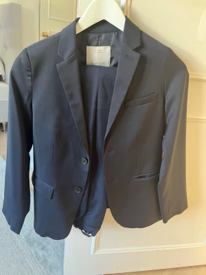 Mörkblå kavaj från Zara Boys och mörkblå kostymbyxor från h&m - Snygg mörkblå kavaj från Zara Boys med klassisk passform. Kavajen har två knappar framtill, två fickor och en bröstficka. Perfekt till mer uppklädda tillfällen och enkel att matcha med skjorta eller t-shirt. Ett par tillhörande kostymbyxor på köpet från h&m, stilrena mörkblå. Storlek 140 cm på både kostymbyxorna och kavajen. Endast använda en gång.