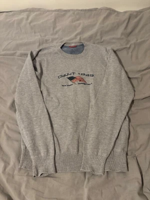Grå sweatshirt från Gant med tryck - Säljer en grå sweatshirt från Gant med broderad amerikansk flagga och texten 'GANT 1949' på bröstet. Tröjan har rund halsringning, ribbade muddar och är gjord i 100% bomull. Perfekt för en avslappnad och stilren look.