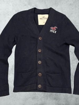 Mörkblå kofta från Hollister - Snygg mörkblå kofta från Hollister med V-ringning och fem bruna knappar framtill. Broderad logga och siffran 1922 på bröstet. Två fickor framtill och ribbade muddar vid ärmslut och nederkant. Tillverkad i mjukt stickat material. Mått: Bredd: 40 cm. Längd: 68 cm. Ärm: 70 cm