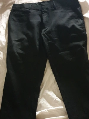 Svarta kostymbyxor med rak passform - Säljer ett par klassiska svarta kostymbyxor med rak passform och bälteshällor. Byxorna har diskreta fickor och stängs med dragkedja och knapp. Perfekta till skjorta eller kavaj för en stilren look. Storlek 30/30