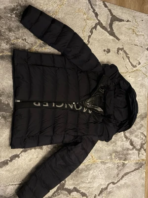 Moncler Jacka - Säljer en svart dunjacka från Moncler med stor logga längs dragkedjan. Jackan har en puffig design, huva och två sidofickor med dragkedja. Perfekt för kalla vinterdagar och riktigt snygg till streetwear-stilen.  OBS FINNS KVITTO TILL JACKAN