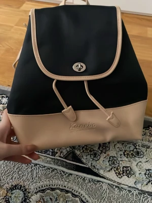 Svart och beige ryggsäck Kanebo - Snygg ryggsäck från Kanebo i svart och beige med dragsko och lock med vridlås. Väskan har beige detaljer och botten i läderimitation samt präglad logga framtill. Perfekt för dig som vill ha en stilren och praktisk accessoar.