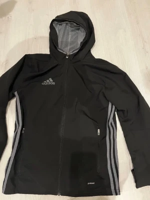 Vindjacka  - Svarat vindjacka från Adidas i storlek XS med huva och hel dragkedja. Jackan har grå ränder längs sidorna, två fickor med dragkedja och meshfoder. Perfekt för träning och aktiva dagar, med ventilerande detaljer på ryggen och snygg sportig look.