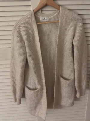 Beige stickad kofta från H&M - Mysig beige stickad kofta från H&M med öppen front och två stora fickor framtill. Koftan har lång ärm och är tillverkad i ett mjukt, ribbat material som passar perfekt för lager på lager. Enkel och stilren design som funkar till det mesta.