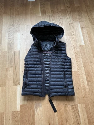 Svart väst från Moose Knuckles - Snygg svart väst från Moose Knuckles med avtagbar huva och quiltad design. Västen har dragkedja framtill och två fickor med dragkedja. Perfekt för streetstyle. Har ett litet hål på ryggen och framsidan som knappt syns och enkelt kan fixas.