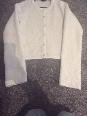 Vit fluffig kofta med pärlknappar - Supersöt vit kofta i fluffigt material med rund hals och pärlknappar framtill. Kortare modell med långa ärmar och ribbade kanter. Perfekt för lager på lager och ger en mjuk, cozy vibe.  Nypris 400 kr