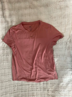 Nike Dri-Fit tränings-tshirt i korall/rosa - Säljer en rosa tränings-tshirt från Nike med klassisk rund hals och diskret Nike-logga på bröstet. T-shirten är kortärmad och tillverkad i ett mjukt, lätt material som passar perfekt för träning och aktiva dagar.