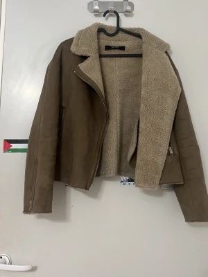 Brun teddyjacka med dragkedja - Snygg brun teddyjacka med mjukt, fluffigt foder och bred krage. Jackan har dragkedja framtill, två sidofickor och långa ärmar. Materialet är fejkmocka på utsidan och teddy på insidan för extra mysfaktor. Perfekt för kalla dagar.