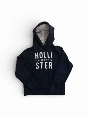 Svart hoodie från Hollister - Mysig svart hoodie från Hollister med stor vit logga på bröstet och texten 'California'. Huvan är fodrad med mjuk, grå teddy för extra komfort. Klassisk känguruficka framtill och ribbade muddar vid ärmar och nederkant. Perfekt för chill dagar.