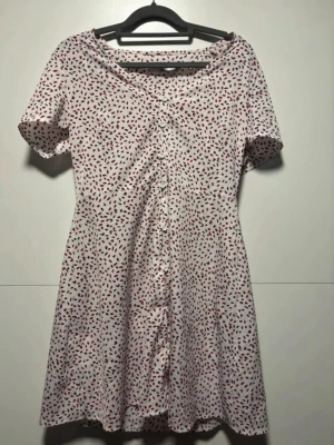 Vit kortklänning med rött mönster - Kortklänning från Wera i vit bas med röda och svarta prickar. Klänningen har korta ärmar, v-ringning och knappar framtill. Materialet känns lätt och passar perfekt för varmare dagar. Snygg och enkel modell som ger en avslappnad men ändå trendig look.