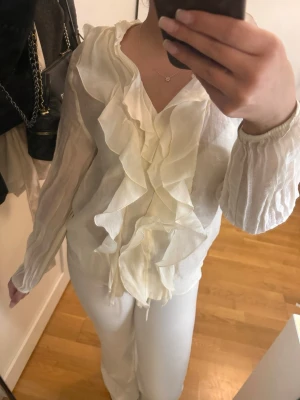 Vit blus med volanger - Hej!💗 Säljer en stilren vit blus med långa ärmar och volangdetaljer längs hela framsidan. Blusen har en djup v-ringning och är tillverkad i ett lätt, något transparent tyg som ger en luftig känsla. Perfekt för en chic och trendig look. Skriv om det är något ni undrar!!🌸