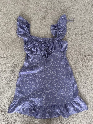 Blommig lila kortklänning med volang - Supersöt lila kortklänning med små vita blommor, volangdetaljer vid axlar och nederkant samt snörning framtill. Klänningen har en romantisk och lekfull stil, perfekt för sommarens alla äventyr. Materialet känns mjukt och lätt.