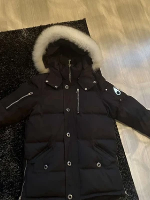 Svart dunjacka från Moose Knuckles - Säljer en svart dunjacka från Moose Knuckles med avtagbar huva och vit fuskpäls. Jackan har flera fickor med dragkedja, silverfärgade knappar och logotyp på ärmen. Perfekt för kalla vinterdagar och riktigt snygg streetstil.