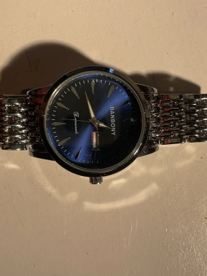 BANBOY klocka med blå urtavla - Stilren klocka från BANBOY med metallarmband i silver och en snygg blå urtavla. Klockan har datumvisning och klassisk rund form. Perfekt accessoar för dig som gillar en elegant look.