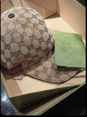 Beige GG keps från Gucci - Säljer en beige keps från Gucci med det klassiska GG-monogrammet över hela kepsen. Kepsen har en böjd skärm och en grön-röd rand på sidan. Tillverkad i bomull och polyester, perfekt för dig som vill ha en ikonisk accessoar.
