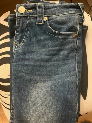 Blå jeans från True Religion - Säljer ett par klassiska blå jeans från True Religion med utsvängda och snygga kontrastsömmar. Jeansen har fem fickor, guldfärgade knappar och detaljerad bakficka med lock och broderi. Tillverkade i slitstark denim med cool tvätt. Har bara använt 2 gånger!!! Köpt på design only i nordstan.