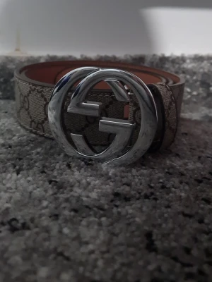 Gucci beige bälte med GG-spänne - Snyggt beige bälte från Gucci med det klassiska GG-mönstret och ett stort, silverfärgat GG-spänne framtill. Bältet har en exklusiv känsla och passar perfekt till jeans eller byxor för att lyfta din outfit.