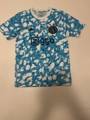Ajax x Adidas blåvit matchtröja - Säljer en Ajax matchtröja från Adidas i blått och vitt med ett unikt kamouflagemönster. Tröjan har Ajax klubbmärke och Ziggo som sponsortryck på bröstet. Tillverkad i lätt och ventilerande material, perfekt för fotboll eller träning.