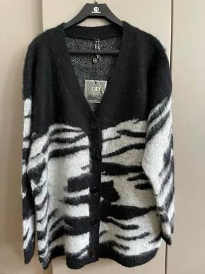 Zebramönstrad kofta från GD - Snygg och mysig kofta från GD med zebramönster i svart och vitt. Koftan har v-ringning, knappar framtill och en loose passform. Perfekt för dig som vill sticka ut med ett djurinspirerat plagg.passar bäst M/L