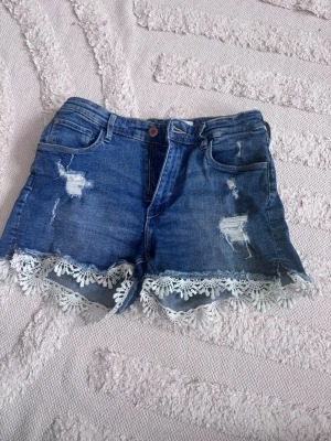 Blå jeansshorts med spetsdetalj - Säljer ett par blå jeansshorts med slitningar och snygg vit spetskant längst ner. Perfekta för sommaren och ger en cool, avslappnad vibe. Ord pris 450kr