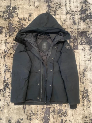 Svart Canada Goose Black label - Snygg svart dunjacka från Canada Goose med stor huva och diskret logotyp på ärmen. Jackan har dragkedja och tryckknappar framtill samt ribbade muddar vid ärmsluten. Perfekt för kalla vinterdagar och har en klassisk, stilren look. Jackan är i storlek S jag är 180cm och 63-67kg passar även i M. Hör gärna av om du har några frågor.🙌