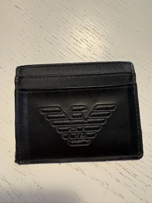 Svart korthållare från Emporio Armani - Snygg svart korthållare från Emporio Armani i läder med präglad logga på framsidan. Smidig och tunn design med flera kortfack både fram och bak. Perfekt för dig som vill ha ett stilrent och exklusivt accessoar i fickan.