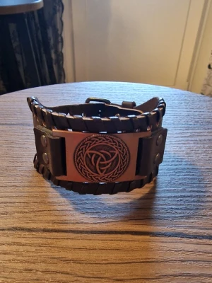 Armband vintage triangel - Svart läderarmband med ett detaljerat triangel i metall. Armbandet har justerbara tryckknappar för att passa olika handledsstorlekar. Storlek 27,5 cm långt. 