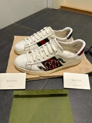 Gucci vita sneakers med orm-motiv - Tja säljer ett par vita sneakers från Gucci med ikoniskt rött och svart orm-motiv på sidan och gröna/röda detaljer. Skorna har vita snören och är tillverkade i läder. Perfekta nu till våren och sommaren 