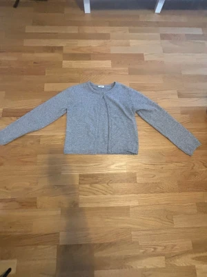 Grå kofta från Kappahl - Mysig grå kofta från Kappahl med rund halsringning och knäppning framtill. Tillverkad i mjukt stickat material och har långa ärmar samt ribbade muddar vid hals och ärmslut. Perfekt att slänga på sig över en t-shirt eller linne.