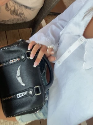 Zadig väska  - Snygg svart handväska i läderimitation med silverfärgade kedjor och ett coolt vingemblem framtill. Väskan har korta handtag och dekorativa nitar längs kanterna. Perfekt accessoar för att lyfta din outfit.