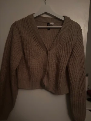 Beige stickad kofta från H&M - Mysig beige stickad kofta från H&M med grov ribbstickning och v-ringad hals. Koftan har två knappar framtill, långa ärmar och en något kortare, boxig passform. Perfekt att slänga på sig över en t-shirt eller linne.