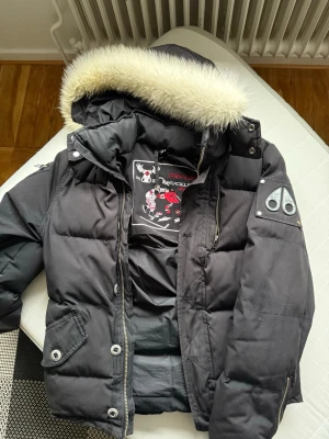 Svart dunjacka Moose Knuckles - Säljer en svart dunjacka från Moose Knuckles med avtagbar huva och snygg pälskant. Jackan har broderad logga på insidan, två framfickor med knappar och dragkedja. Perfekt för kalla vinterdagar och riktigt varm.