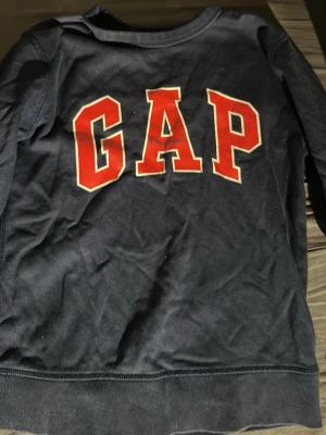 Mörkblå sweatshirt från GAP Kids - Mörkblå sweatshirt från GAP Kids med stora röda bokstäver och vit kant på bröstet. Klassisk rund halsringning och mjukt material som passar perfekt till vardagslooken. Snygg och enkel design med ribbade muddar.