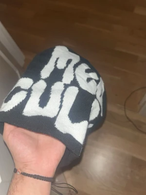 Svart mössa med vit text 'Mea Culpa' - Säljer en svart mössa med stor vit text där det står 'Mea Culpa' i ett coolt, grafiskt typsnitt. Mössan är stickad och har en avslappnad passform som passar perfekt till streetwear-stilen. Materialet känns mjukt och bekvämt mot huvudet.