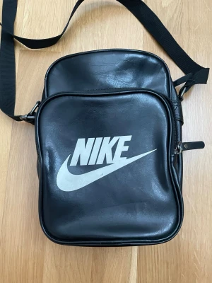 Svart axelväska från Nike - Snygg svart axelväska från Nike med stort vitt logotryck på framsidan. Väskan har justerbar axelrem, två fack med dragkedja och är tillverkad i ett glansigt konstlädermaterial. Perfekt för att bära det viktigaste och ger en sportig vibe.