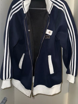 Marinblå hoodie med vita detaljer - Snygg marinblå hoodie från Hammarby med vita muddar, dragkedja och klassiska vita ränder längs ärmarna. Två fickor framtill och en liten bröstdetalj med knapp. Tillverkad i mjukt sweatshirtmaterial, perfekt för en avslappnad streetstil.