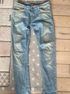 Ljusblå jeans från Nudie Jeans skinny lin  - Säljer ett par ljusblå jeans från Nudie Jeans med klassisk femficksmodell och orangea sömmar. Jeansen har en slim passform och snygga slitningar framtill och bak, samt Nudie-logga på bakfickan. Tillverkade i bomull för en skön känsla.