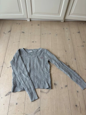 Grå kabelstickad tröja Hollister - Snygg grå kabelstickad tröja från Hollister med V-ringning och diskret logga på bröstet. Tröjan har långa ärmar och ribbade muddar vid ärmslut och nederkant. Perfekt för lager på lager och en chill vardagslook.