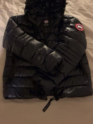 Canada Goose Crofton - Snygg  dunjacka från Canada Goose med huva och klassisk logga på vänster ärm. Jackan har glansigt material, dragkedja framtill och justerbar nederkant. Perfekt för kalla vinterdagar och riktigt varm tack vare dunfyllningen.