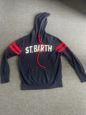 Marinblå hoodie St. Barth - Snygg marinblå hoodie från St. Barth med vita bokstäver på bröstet och röda ränder på ärmarna. Hoodien har röda dragsnören och en stor ficka framtill. Perfekt för en avslappnad och sportig stil. Fler frågor eller funderingar hör av er i Dm!
