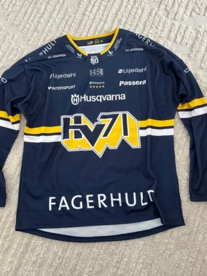 HV71 hockeytröja blå/gul Bauer - Säljer en blå HV71 hockeytröja från Bauer med gula och vita detaljer. Tröjan har lång ärm, tryckta sponsorer och stort HV71-märke på bröstet. Tillverkad i polyester, perfekt för hockeyfans eller till matchen. Klassisk sportig look med rund hals.