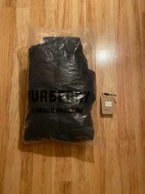 Svart pufferjacka från Burberry - Snygg svart pufferjacka från Burberry med huva och dragkedja framtill. Jackan har två fickor med dragkedja och diskret Burberry-logga på ärmen. Perfekt för kalla vinterdagar och riktigt trendig stil. Passar även till våren eftersom man kan dra av ärmarna på jackan. Pufferjackan passar perfekt för både vintern och våren.