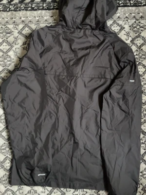 Svart vindjacka från Nike med huva - Snygg svart vindjacka från Nike med dragkedja framtill och justerbar huva. Jackan har flera reflekterande detaljer och Nike-logga på bröstet. Tillverkad i lätt och tåligt material som skyddar mot vind. Perfekt för utomhusaktiviteter och streetwear.