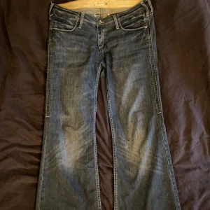 Mörkblå jeans med bootcut - Säljer ett par snygga mörkblå jeans i storlek 40. De har en klassisk bootcut-stil och är perfekta för en avslappnad look. Jeansen har en knapp och dragkedja framtill samt fickor både fram och bak. De är i bra skick med lite slitage vid fållen, vilket ger dem en cool vintage-känsla.