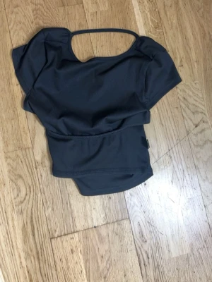 Svart croppad topp med öppen rygg - Trendig svart croppad topp med unik öppen rygg och korta ärmar. Toppen har en smal rem över ryggen som ger en cool detalj. Perfekt att styla till jeans eller kjol för en modern look.