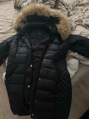 Rockandblue jacka  - Snygg svart lång dunjacka från rockandblue med quiltade detaljer och stor huva med fuskpäls. Jackan har dragkedja och knappar framtill samt två snedställda fickor med dragkedja. Perfekt för kalla vinterdagar.