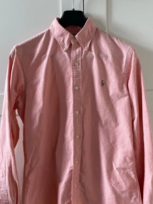 Rosa skjorta från Ralph Lauren - Klassisk rosa skjorta från Ralph Lauren med button-down krage och broderad logga på bröstet. Skjortan har långa ärmar, normal passform och stängs med vita knappar framtill. Tillverkad i mjuk bomull som känns skön mot huden.