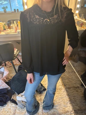 Svart blus med spetsdetaljer - Snygg svart blus med lång ärm och vackra spetsdetaljer upptill vid halsen och axlarna. Blusen har en lös passform och rund halsringning. Perfekt att styla med jeans eller kjol för en trendig look.